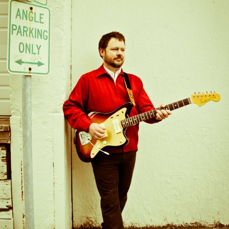 Man standing with a guitar in front of a peeling painted wall