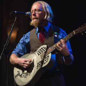 Jason Reichart, a man with long sandy hair, plays a guitar on a dark stage