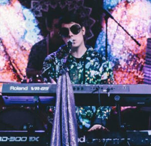 Photo of man in sunglasses playing a keyboard in front of a glitter backdrop